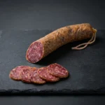 Salchichón Bellota Arturo Sánchez u komadu, s vidljivim komadićima mesa i bijele masnoće na presjeku i narezane šnite.