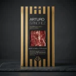Arturo Sánchez pršut Bellota 100 % Ibérico u crno-zlatnom pakiranju s vidljivim šnitama.